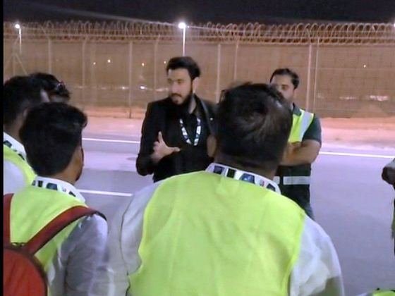 Ibrahim briefing a crew in hi-vis on an airside apron at night, perimeter fence behind